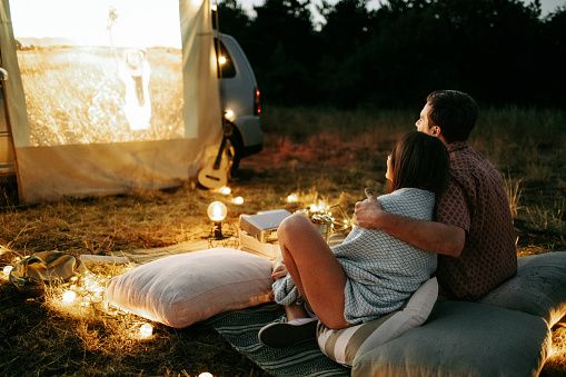Coppia giovane seduta davanti a un'auto durante una proiezione di cinema all'aperto, a illustrare un incontro serio, la comunicazione e i primi momenti di una relazione duratura.