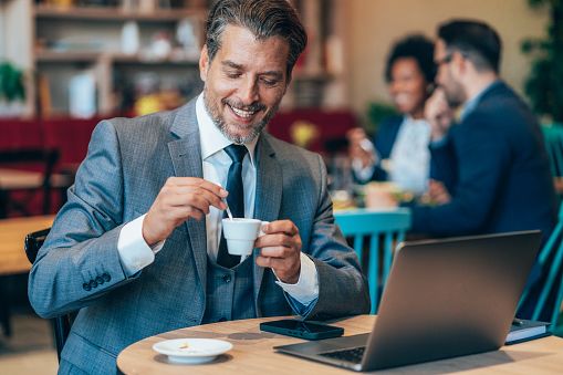 Uomo sulla quarantina sorridente in un caffè con un laptop e una tazza di caffè, che illustra un incontro serio, una prima impressione positiva e una comunicazione autentica.
