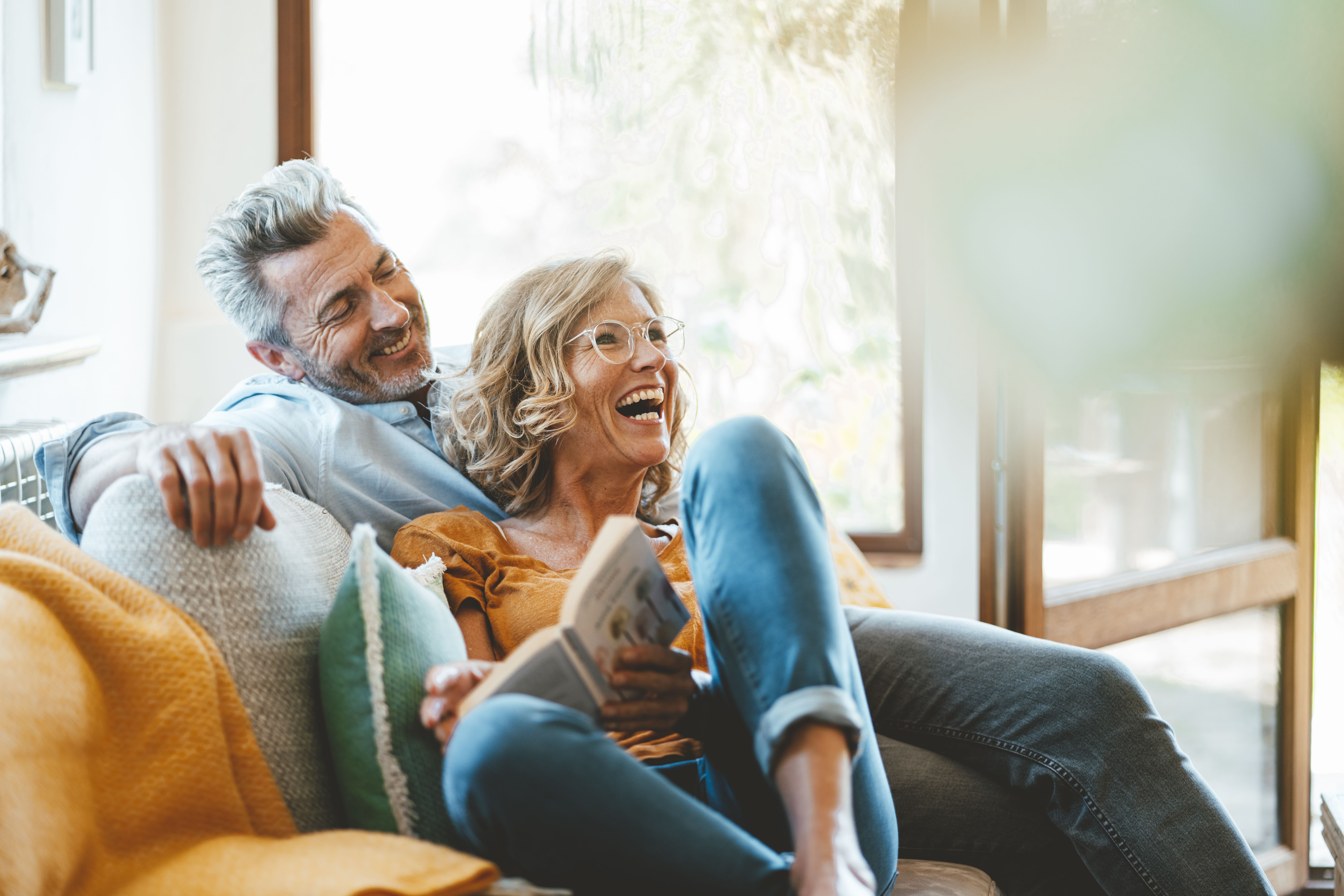 Coppia sulla quarantina sorridente su un divano a casa, che condivide un momento di complicità, a illustrare una relazione duratura, valori comuni e un incontro serio riuscito.