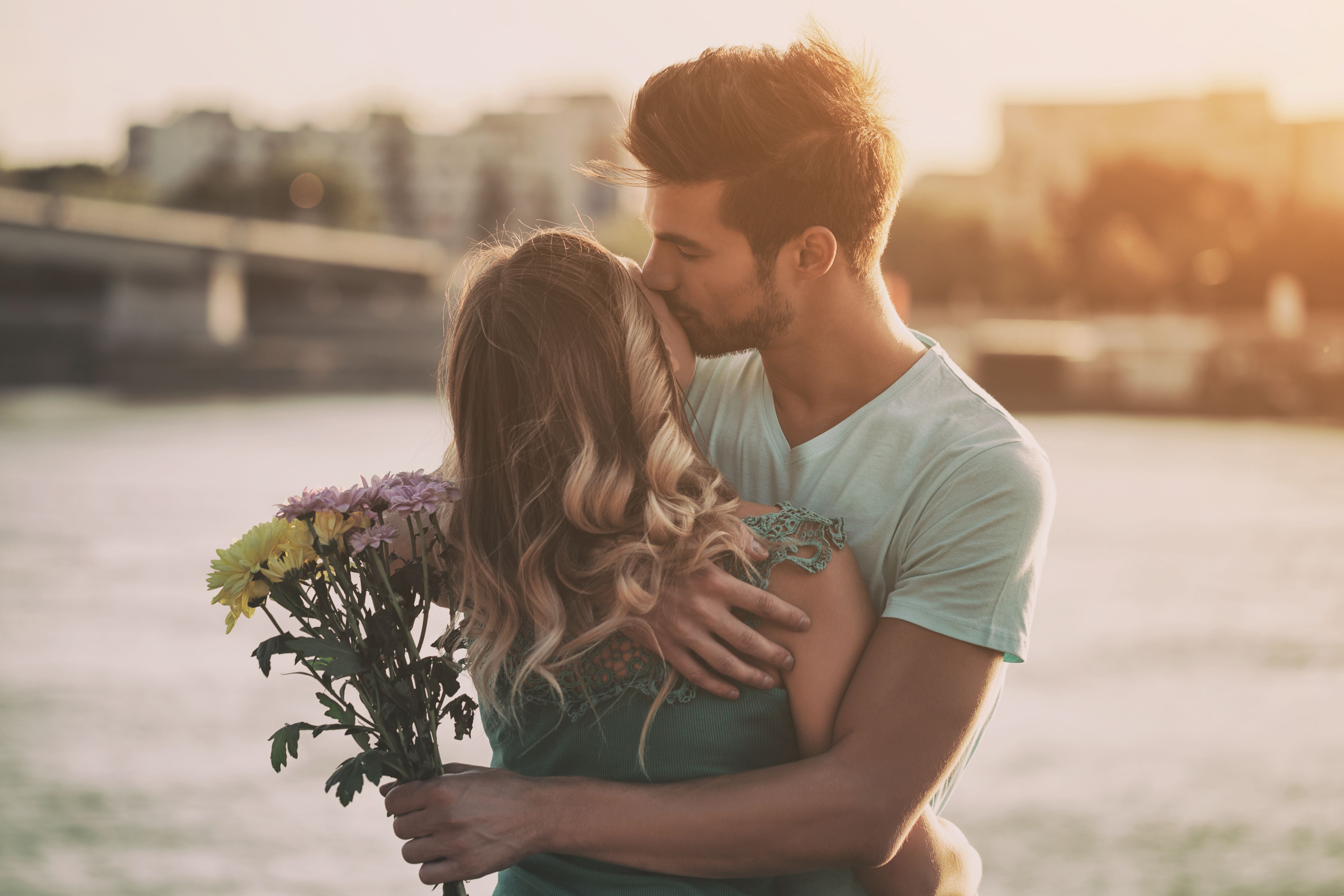 Giovane coppia sorridente che si abbraccia in città, con l’uomo che tiene un mazzo di fiori, a illustrare un incontro serio, una relazione duratura e un primo appuntamento riuscito.